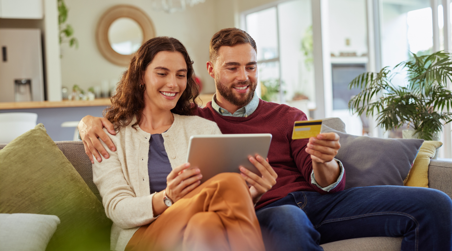 Young couple holding iPad and debit card together on couch, smiling.