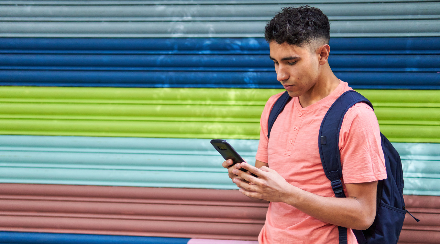 Male student against colored wall with backpack, checking mobile phone.